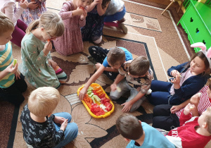 Zdjęcie przedstawia grupę dzieci siedzących na dywanie i jedzących owoce i warzywa.