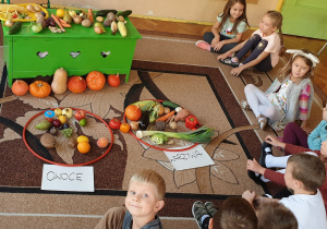 Na zdjęciu dzieci siedzą na dywanie i dzielą przyniesione owoce i warzywa na odpowiednie kategorie.