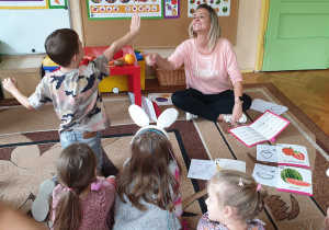 Zdjęcie przedstawia nauczycielkę i dzieci siedzące na dywanie i chłopca zgłaszającego się do odpowiedzi.