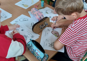 Zdjęcie przedstawia dzieci siedzące przy stoliku i kolorujące rysunki owoców.