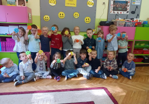 Na zdjęciu dzieci z grupy Skrzaty pozują do zdjęcia grupowego, uśmiechają się i trzymają kolorowe uśmiechnięte buźki.