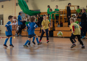 Zdjęcie przedstawia chłopców w niebieskich i żółtych strojach rozgrywających mecz na hali sportowej.