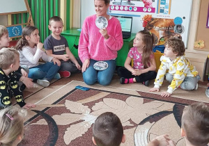 Na zdjęciu dzieci siedzą na dywanie i obserwują jak nauczycielka pokazuje i tłumaczy schemat powstawania zaćmienia.