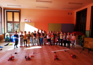 Grupa dzieci stoi w półkolu , przed dziećmi na podłodze szereg małych drukarek 3D.