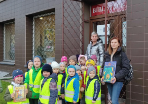 Panie i dzieci w kamizelkach odblaskowych stojące przed budynkiem Biblioteki.