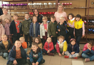 Na zdjęciu grupa razem z paniami pozuje na tle regałów z gotowymi bombkami.