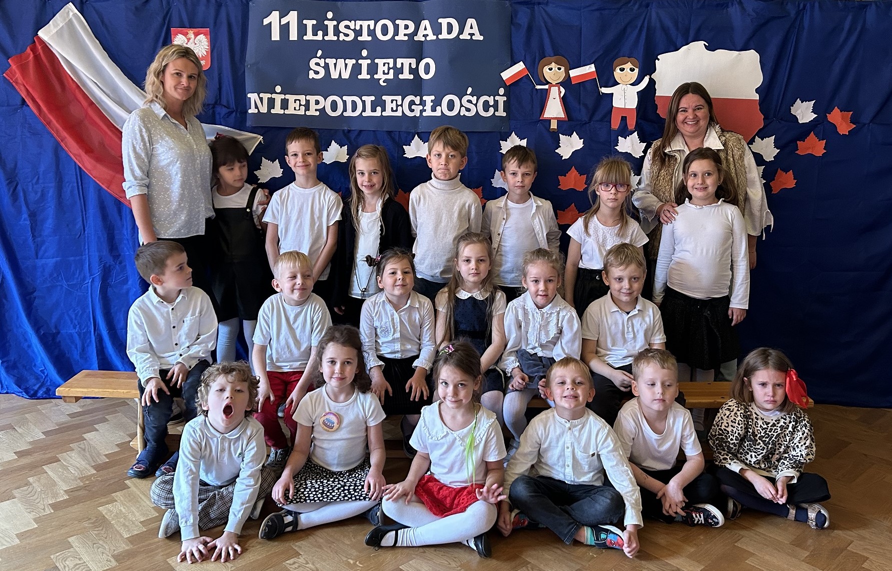 Grupa dzieci z nauczycielkami w galowych strojach pozuje na tle napisu Święto Niepodległości i flagi Polski.