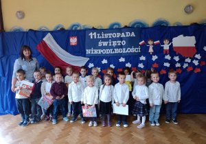 Dzieci z grupy drugiej wraz z nauczycielką pozują w odświętnych strojach na tle dekoracji z okazji 11 listopada