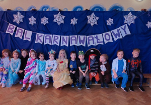 Dzieci w strojach karnawałowych siedzące na ławce. Zdjęcie grupowe na tle dekoracji karnawałowej.