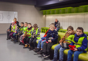 dzieci siedzą na ławkach w szatni dla piłkarzy, słuchają prowadzącego o historii stadionu i o grających dawniej i dziś piłkarzach