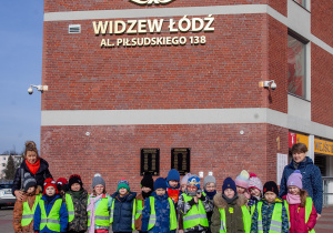 grupa dzieci stoi pod stadionem, w tle na ścianie stadionu widać wielki znaczek stadionu Widzewa