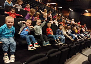 Na zdjęciu dzieci siedzą uśmiechnięte na widowni w teatrze, czekając na przedstawienie.