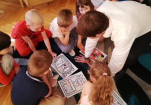 Grupa dzieci siedzi na dywanie, przed sobą dzieci trzymają rysunki z plecaczkami, wśród dzieci siedzi wiceprezydent Łodzi, który omawia z dziećmi ich rysunki.