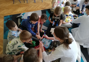 Dzieci siedzą na dywanie i pod kierunkiem studenta medycyny uczą się używając zabawkowych narzędzi medycznych badać swoje maskotki misiów, które są ich pacjentami.