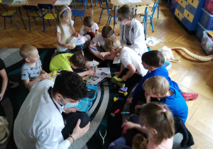 Dzieci siedzą na dywanie i pod kierunkiem studenta medycyny uczą się używając zabawkowych narzędzi medycznych badać swoje maskotki misiów, które są ich pacjentami.