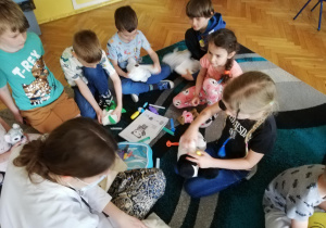 Dzieci siedzą na dywanie i pod kierunkiem studenta medycyny uczą się używając zabawkowych narzędzi medycznych badać swoje maskotki misiów, które są ich pacjentami.