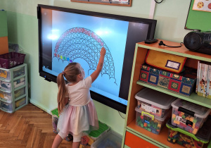 Dzieci kolorują kropkową tęczę na tablicy multimedialnej.