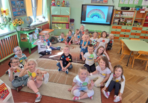 Dzieci uśmiechają się do zdjęcia, w tle obraz kropkowej tęczy, pokolorowany przez dzieci na tablicy multimedialnej