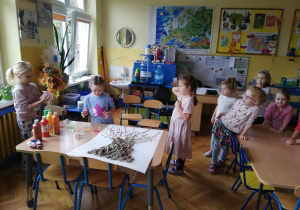 dzieci stoją przy stoliku, na którym leży karton z pracą plastyczną - drzewem i czekają w kolejce na stemplowanie paluszkami na kolorowych liści farbą na drzewie, część dzieci już stempluje umoczonymi w kolorowej farbie paluszkami jesienne liście