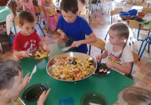 .dzieci przy stoliku, wrzucają do dużej misy pokrojone przez siebie owoce