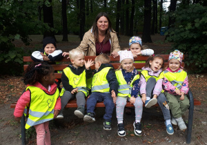 zdjęcie przedstawia dzieci wraz z nauczycielką siedzące na parkowej ławeczce i stojące przy niej.