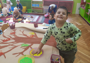 na zdjęciu chłopiec podczas wykonywania pracy plastycznej ,,Drzewo” w tle dzieci podczas zabawy swobodnej na dywanie