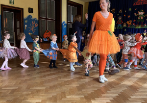Na zdjęciu dzieci w jesiennych przebraniach idą ustawione "w pociąg" za nauczycielką ubraną na pomarańczowo