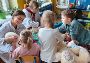 -na zdjęciach dzieci przy przedszkolnych stolikach bawią się wraz ze studentami medycyny w lekarzy i pacjentów. wykorzystują do tego zabawki - pluszaki przyniesione przez dzieci oraz zabawkowy i prawdziwy sprzęt medyczny: plasterki, bandaże, strzykawki, stetoskopy, a także oglądają zdjęcia rtg. i podają na niby syropki
