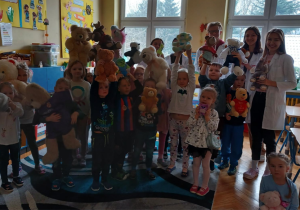 na zdjęciu grupa dzieci stoi trzymając w rękach swoje zabawki - pluszaki, za dziećmi stoją studenci medycyny , ubrani w białe , lekarskie fartuchy.