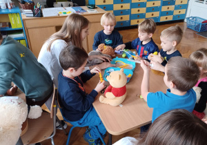 -na zdjęciach dzieci przy przedszkolnych stolikach bawią się wraz ze studentami medycyny w lekarzy i pacjentów. wykorzystują do tego zabawki - pluszaki przyniesione przez dzieci oraz zabawkowy i prawdziwy sprzęt medyczny: plasterki, bandaże, strzykawki, stetoskopy, a także oglądają zdjęcia rtg. i podają na niby syropki