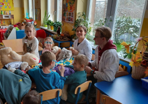 -na zdjęciach dzieci przy przedszkolnych stolikach bawią się wraz ze studentami medycyny w lekarzy i pacjentów. wykorzystują do tego zabawki - pluszaki przyniesione przez dzieci oraz zabawkowy i prawdziwy sprzęt medyczny: plasterki, bandaże, strzykawki, stetoskopy, a także oglądają zdjęcia rtg. i podają na niby syropki