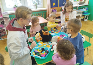 Dzieci siedzące przy stolikach wykonują przykładowe zabiegi lekarskie na swoich pluszakach