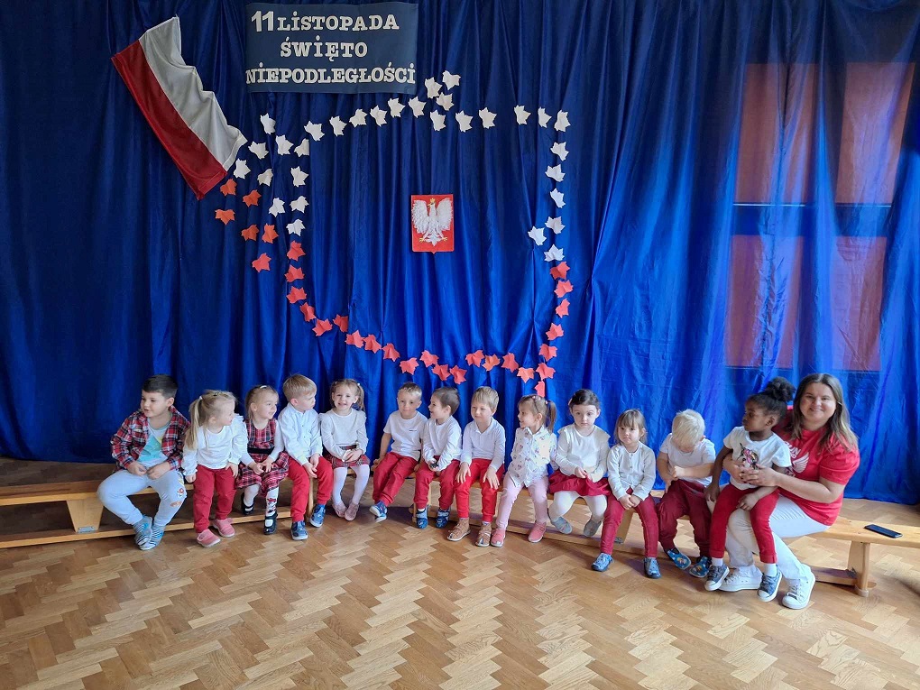 zdjęcie przedstawia dzieci z grupy Misie wraz z nauczycielką podczas uroczystości z okazji Narodowego Święta Niepodległości na sali gimnastycznej
