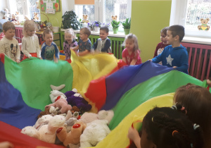 Zdjęcie przedstawia dzieci podczas zabawy ruchowej przy muzyce z wykorzystaniem chusty animacyjnej i pluszaków