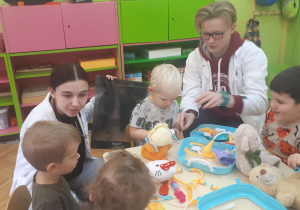 zdjęcie przedstawia dzieci i studentów siedzące przy stolikach podczas zabawy z wykorzystaniem pluszowych maskotek i zabawkowych akcesorii medycznych