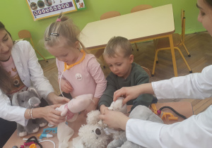 zdjęcie przedstawia dzieci i studentów siedzące przy stolikach podczas zabawy z wykorzystaniem pluszowych maskotek i zabawkowych akcesorii medycznych