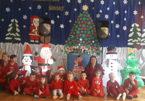 zdjęcie przedstawia dzieci z grupy ,, Misie” wraz z Paniami podczas balu mikołajkowego siedzące na Sali gimnastycznej na tle dekoracji