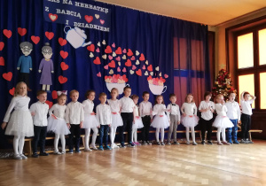 Dzieci stojące obok siebie w tle dekoracja z serduszek dla babć i dziadków.