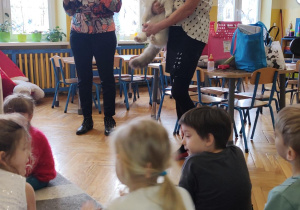 Stojce dwie panie, jedna trzymająca kota i dzieci siedzące na dywanie.