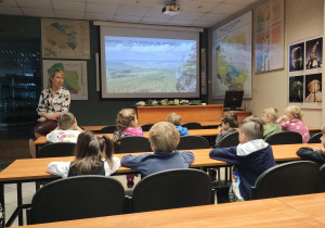 Dzieci siedzące w ławkach podczas prezentacji edukacyjnej.