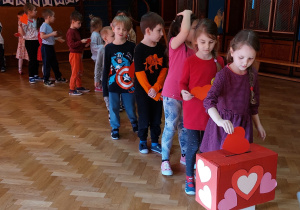 1. Dzieci stoją z kartkami walentynkowymi w rzędzie, po kolei wrzucają kartki do pudełka ozdobionego serduszkami - walentynkowej skrzynki