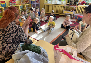 Na zdjęciu dzieci siedzą na dywanie i słuchają wolontariuszek z Fundacji Kocia Mama opowiadających o kotach.