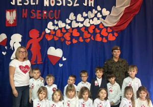 Na zdjęciu grupa Pszczółek z nauczycielką w mundurze żołnierskim na tle napisu patriotycznego.