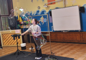 Na zdjęciu Pani za pomocą specjalnego sprzętu tworzy gigantyczne bańki mydlane.