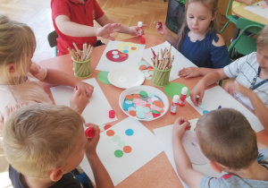 Zdjęcie przedstawia czworo dzieci siedzących przy stoliku przyklejające kolorowe kropki na białym kartonie.