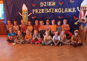 Zdjęcie przedstawia dzieci w pomarańczowych strojach na tle granatowego materiału z napisem "Dzień przedszkolaka"