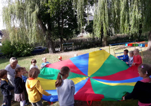 Dzieci z grupy piątej wraz z nauczycielką podczas zabawy w ogrodzie kolorową chustą