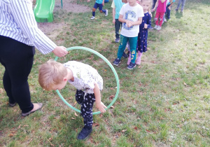 na zdjęciu nauczycielka trzyma koło hoola-hop, przez środek którego przechodzi chłopiec, w tle dzieci ustawione w rządek, które patrząc na kolegę czekają na swoja kolej