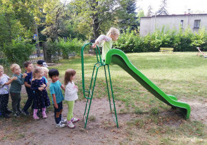 na zdjęciu widać zjeżdżalnię w ogródku przedszkolnym, na której dziewczynka szykuje się do zjazdu oraz dzieci czekające na swoją kolej.