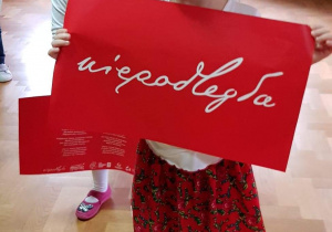 Dziewczynka ubrana w strój ludowy trzyma napis "Niepodległa"